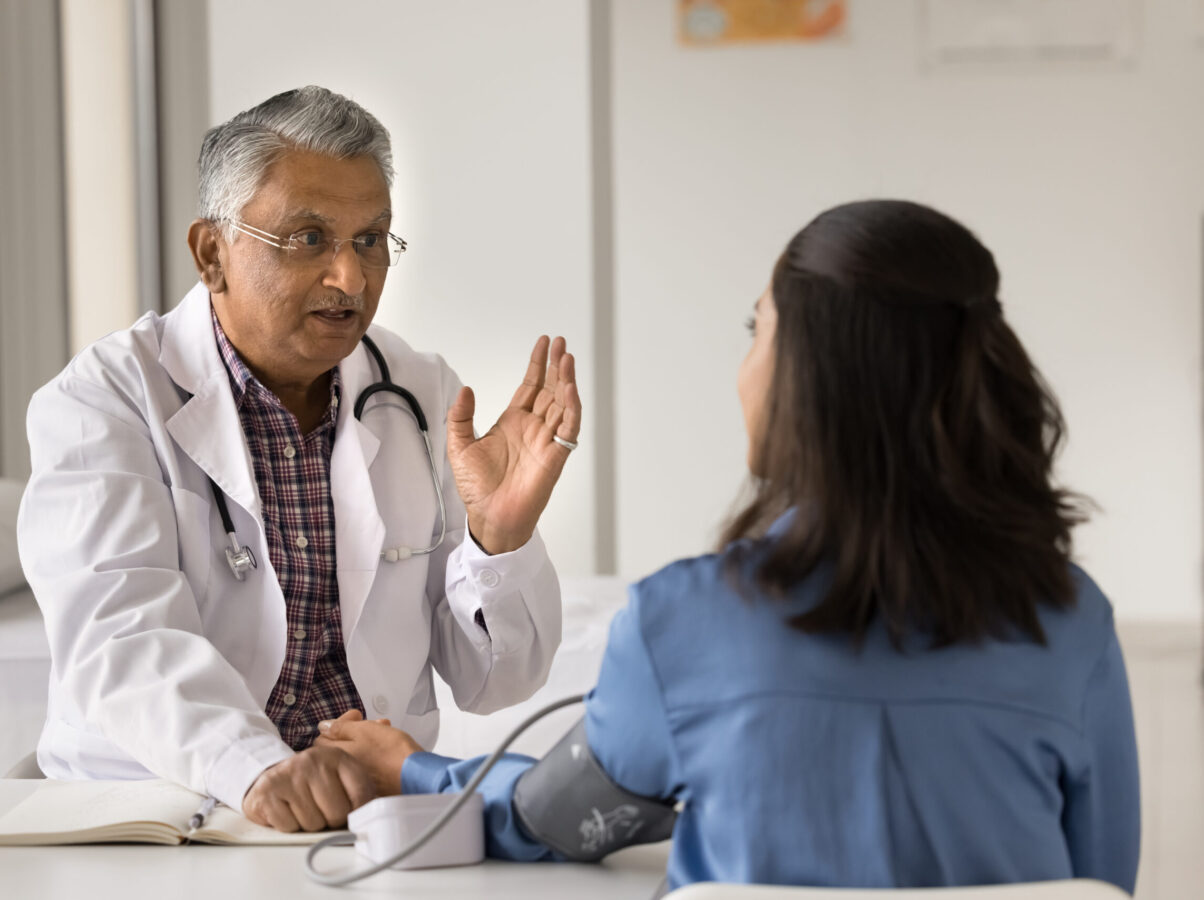 Older Indian doctor man examining young patient with complaints on hypertension, measuring blood pressure, talking to woman, discussing healthcare checkup, examination